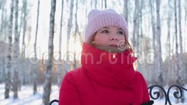 穿着红色温暖夹克的年轻美女坐在<strong>画报</strong>公园的长凳上，在冬天寒冷的阳光明媚的日子里，用桦树和温暖的双手